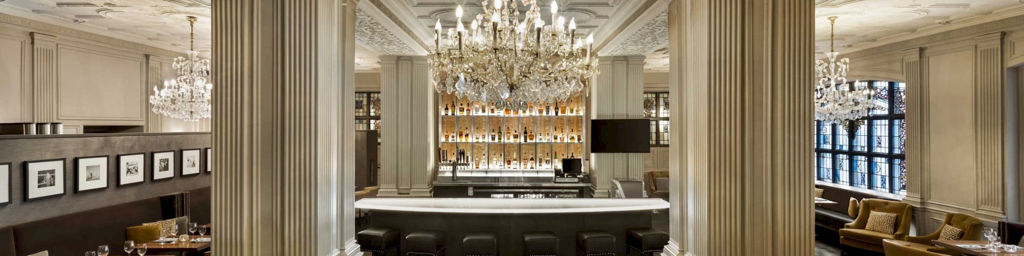 A spacious, elegant bar area with chandeliers, white columns, a well-stocked counter, and cozy seating booths around the perimeter, exuding a classic ambiance.