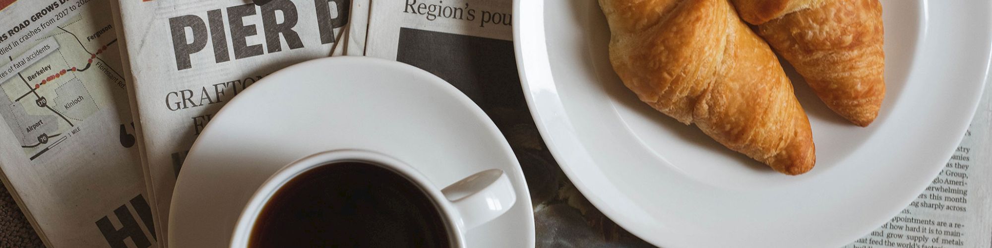 A breakfast scene with two croissants on a white plate, a cup of coffee on a saucer, newspapers, and a business card from 