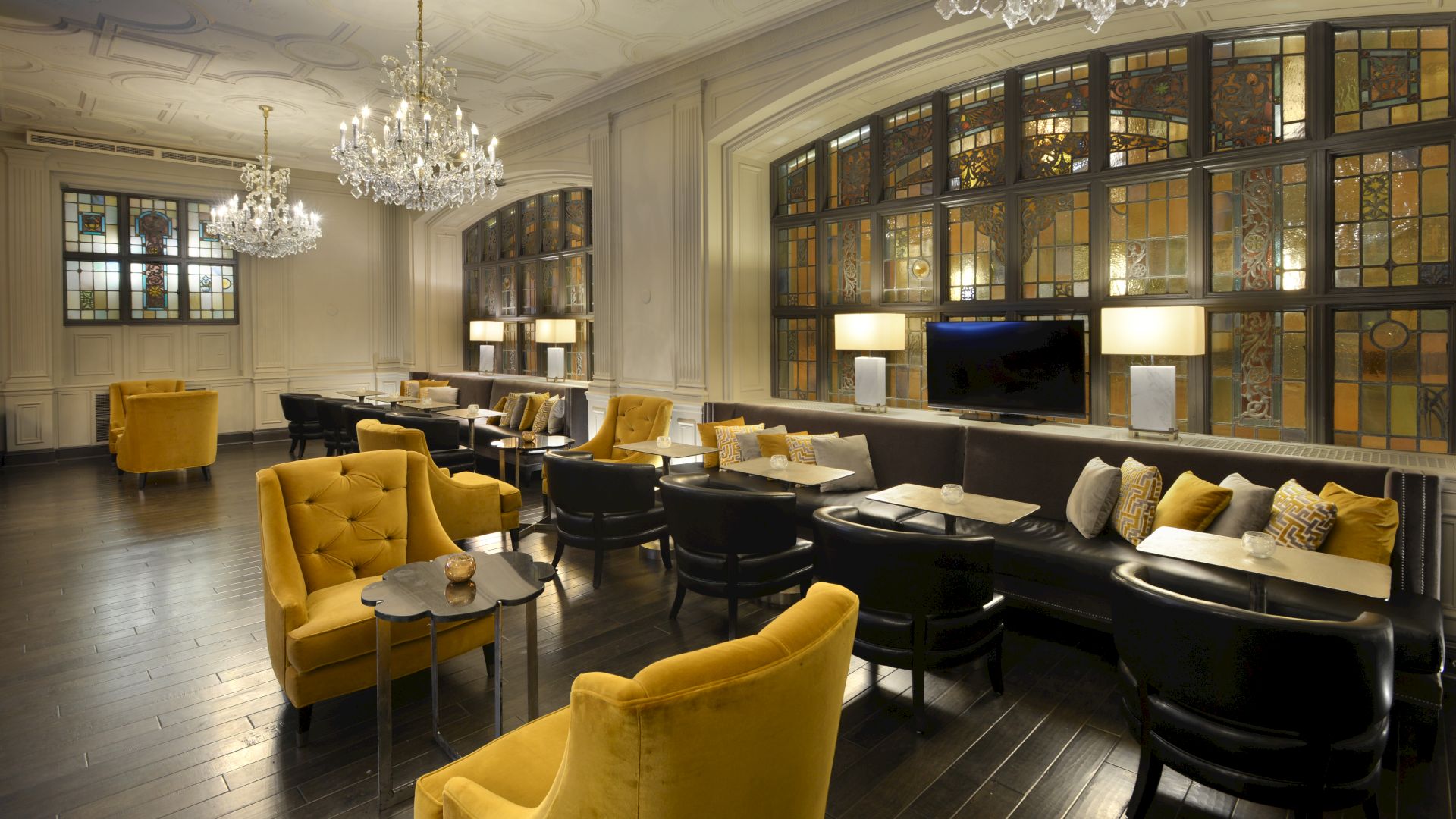 A stylish lounge with yellow armchairs, chandeliers, and large windows, containing tables and a cozy seating arrangement, is seen in the image.