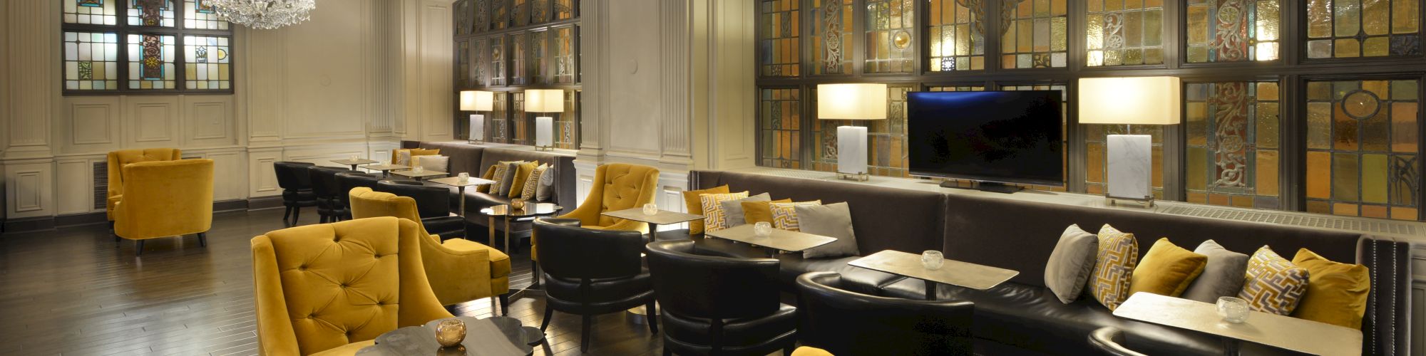 A stylish lounge with yellow armchairs, chandeliers, and large windows, containing tables and a cozy seating arrangement, is seen in the image.