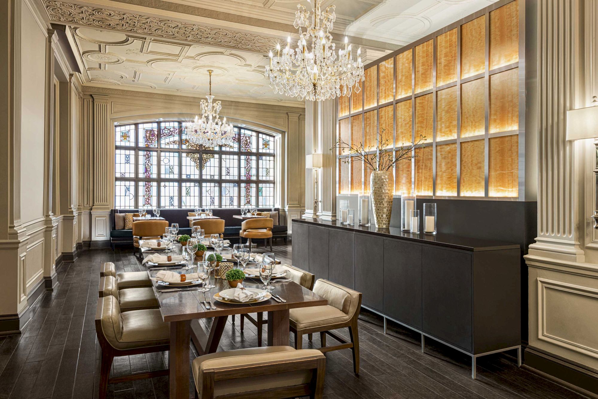 A well-lit, elegant dining room with chandeliers, wooden table, chairs, large window, decorative wall paneling, and set dining table.