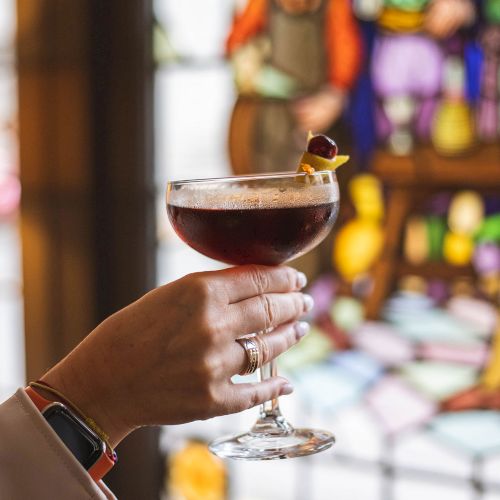 A hand holding a cocktail glass with a reddish drink garnished with a cherry and lemon twist, in front of a colorful stained glass window.