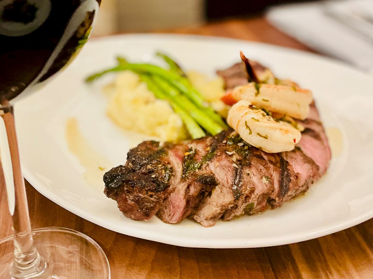 A plate of steak topped with shrimp, served with mashed potatoes and asparagus, alongside a glass of red wine on a wooden table.