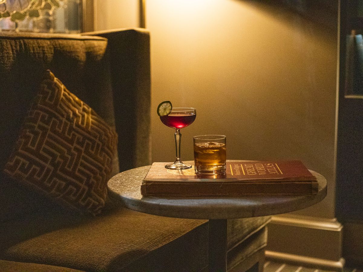 A cozy setting with a sofa and patterned cushion, a round table with two drinks and a book, illuminated by warm light, creating a calm ambiance.