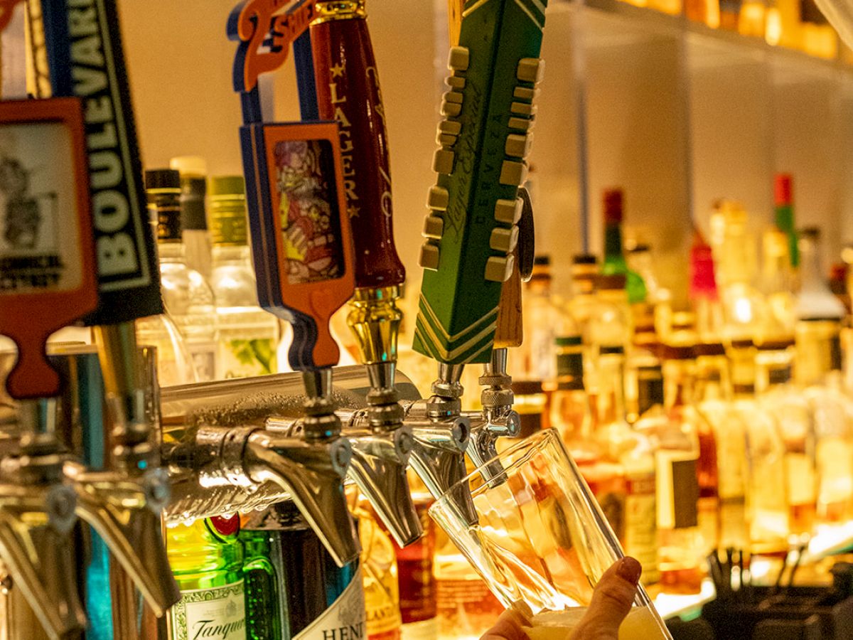 A bartender is pouring a drink from a tap in a well-stocked bar, with various bottles and tap handles visible in the background.