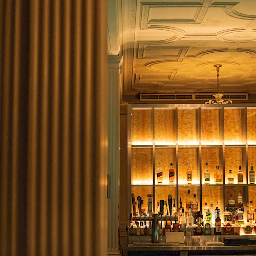 A dimly lit bar with a wooden backlit shelf full of bottles, warm tones, and a ceiling fan above; cozy nightlife ambiance.