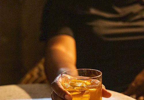 A person in a dark shirt sits at a table, holding a glass of amber liquor with ice, reaching forward.