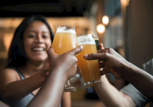 A group of people clinking beer glasses together in a toast, smiling and enjoying a casual gathering indoors.