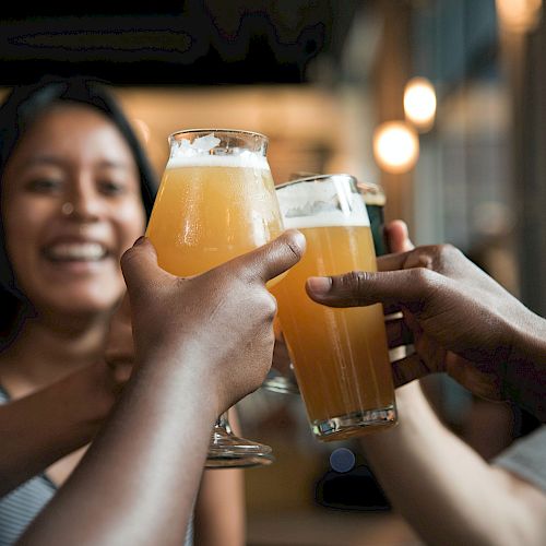 A group of people clinking beer glasses together in a toast, smiling and enjoying a casual gathering indoors.