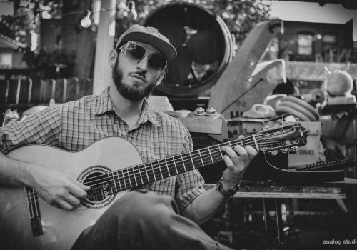 A man with sunglasses and a hat plays an acoustic guitar outdoors, surrounded by various objects and instruments. The photo is in black and white.