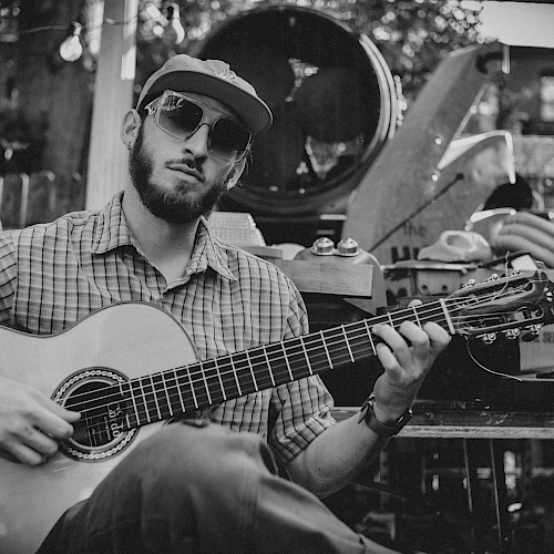 A man with sunglasses and a hat plays an acoustic guitar outdoors, surrounded by various objects and instruments. The photo is in black and white.