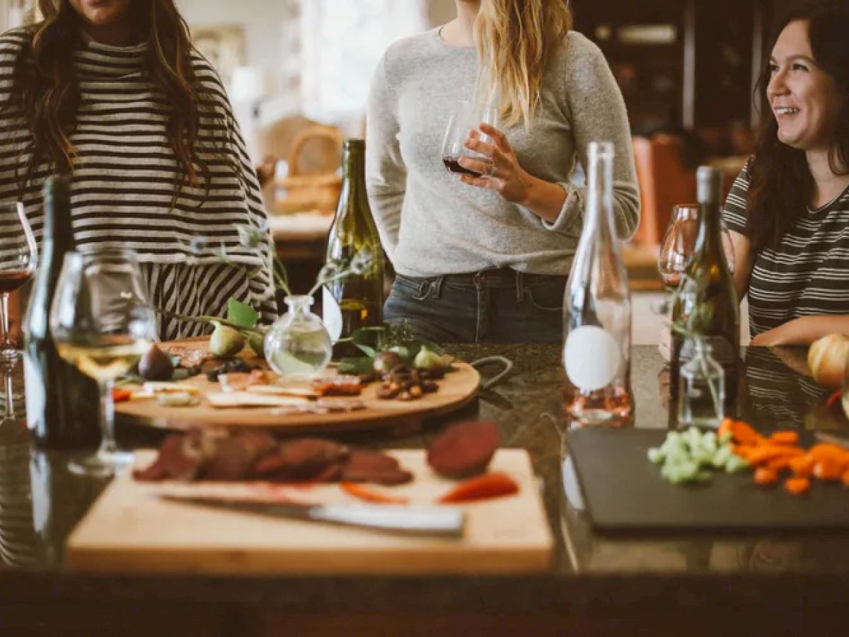 People are gathered around a countertop with wine glasses and food, including charcuterie and vegetables, enjoying a casual get-together.