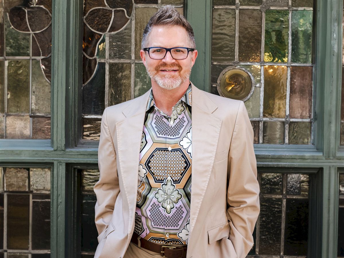 A man in a beige suit and patterned shirt stands in front of a vintage window with decorative panes and hands in his pockets, smiling at the camera.