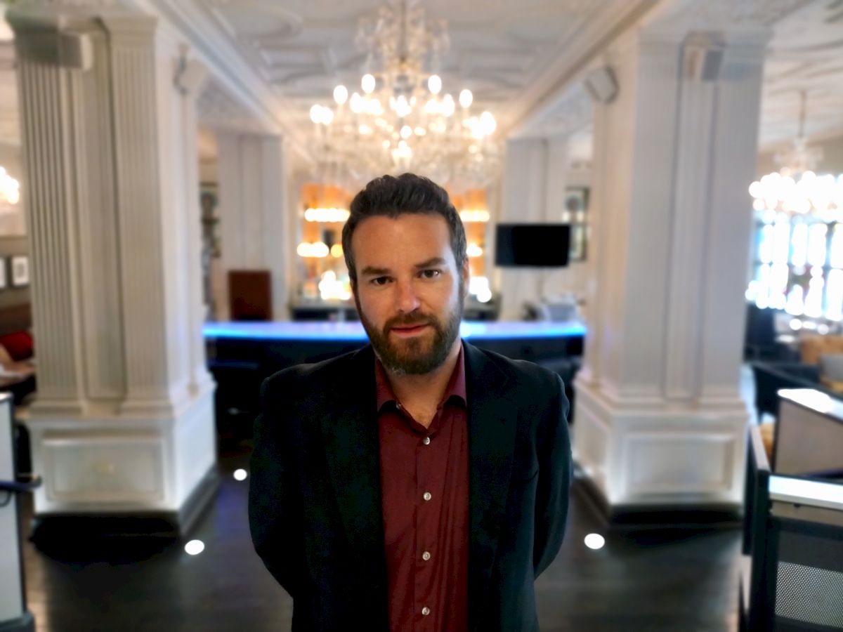 A man is standing in a well-lit, elegant room with chandeliers, white pillars, and a bar area in the background, wearing a dark blazer and red shirt.