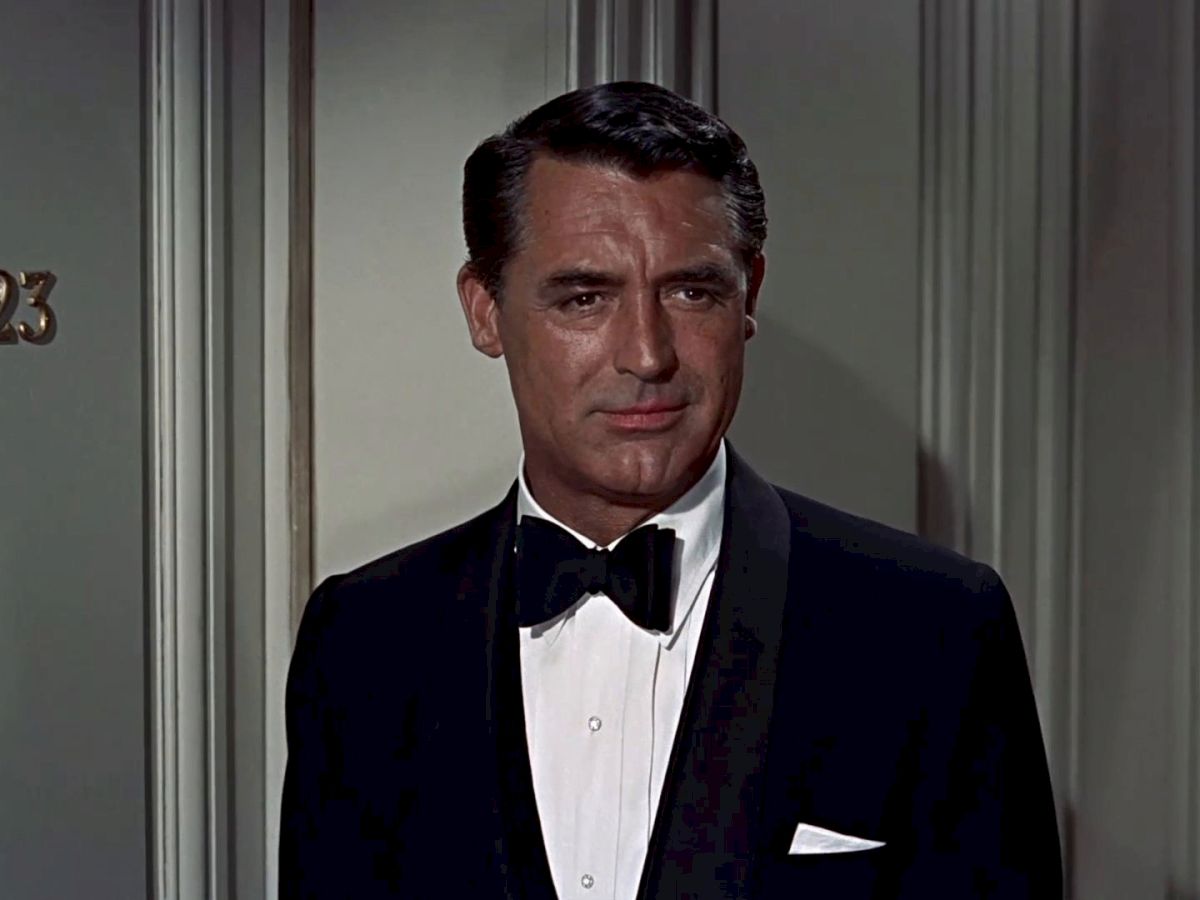 A man in a black tuxedo stands in front of a door numbered 623, wearing a white shirt and black bow tie.