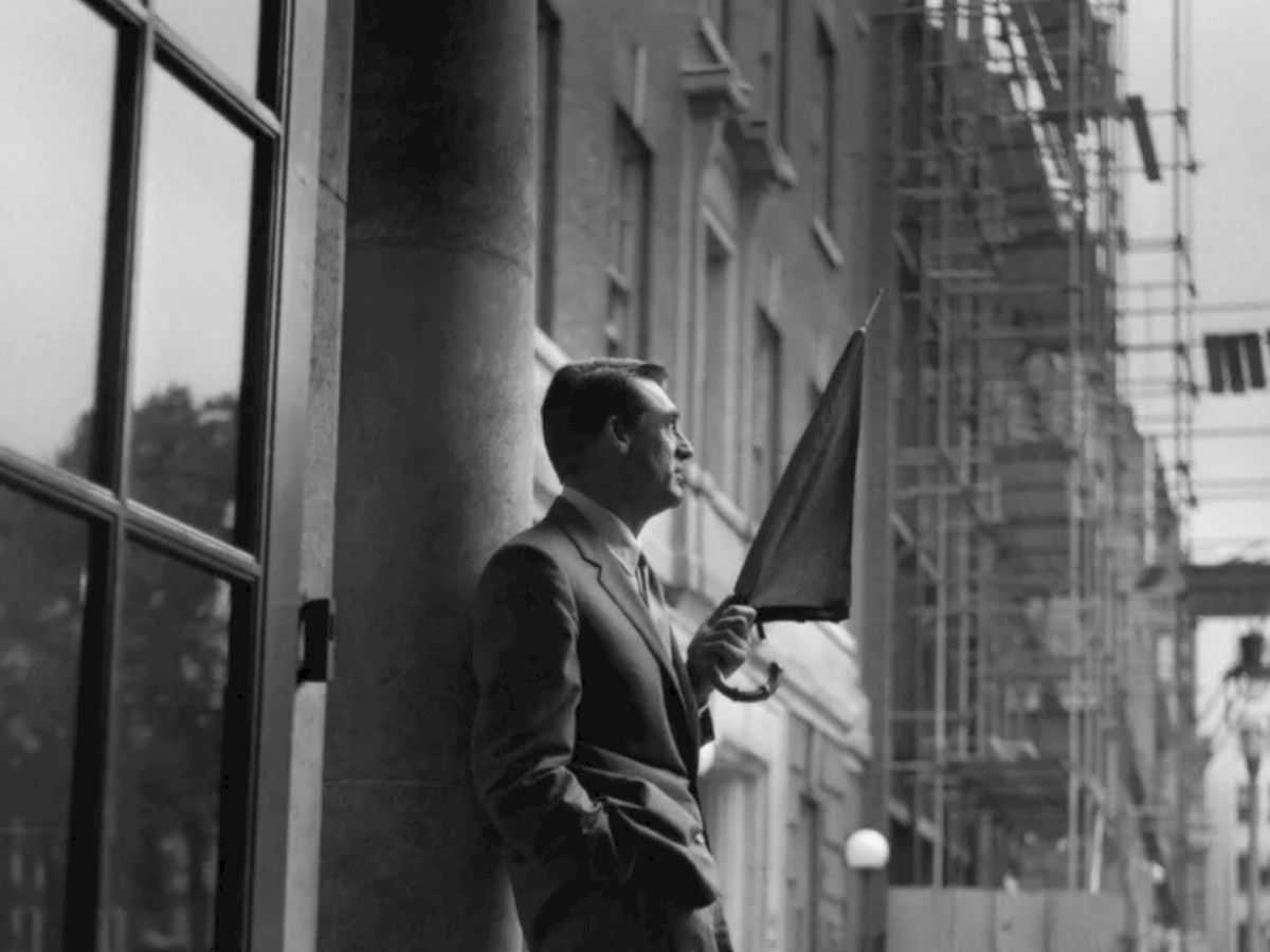 The image shows a man in a suit holding an umbrella, standing against a building column. There is scaffolding in the background and a city street scene.