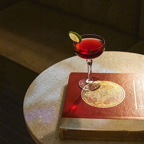 A cocktail glass with a red drink garnished with a lime slice sits on a book on a round table, with a cushioned seat in the background.