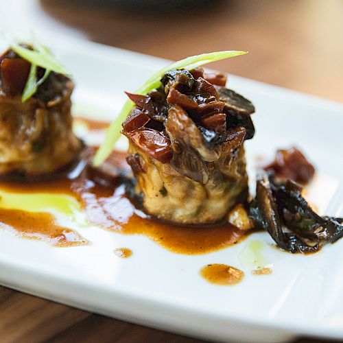 The image shows a plated dish with two small servings of food topped with garnishes and sauce, presented on a white plate.