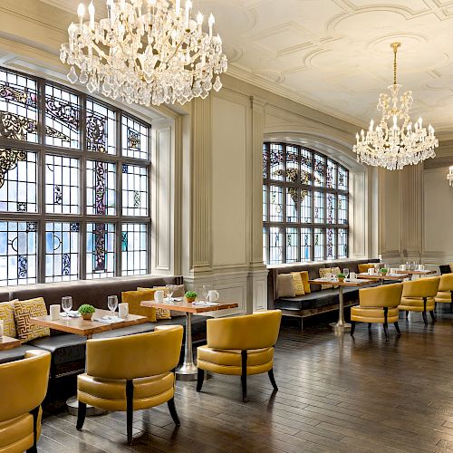 An elegant dining room with yellow chairs, chandeliers, and large stained glass windows, featuring neatly set tables and a polished wooden floor.