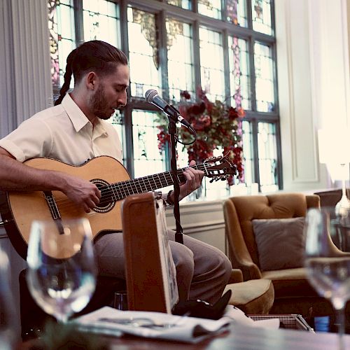 A man is seated and playing an acoustic guitar while singing into a microphone in a cozy room with tables, chairs, and decorative lights.