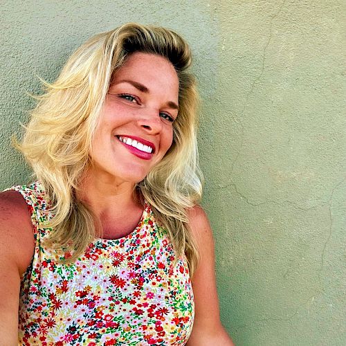 The image shows a smiling person with blonde hair, wearing a floral-patterned top, standing against a plain beige wall.