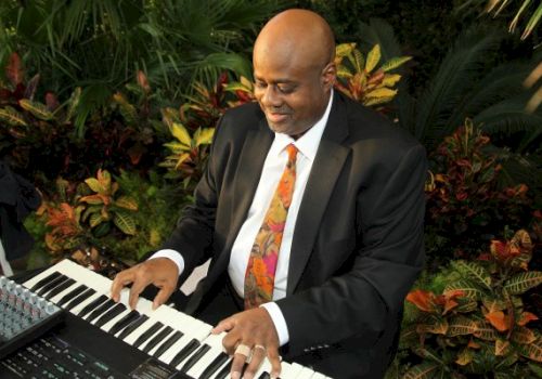 A man in a black suit plays a keyboard surrounded by lush greenery, appearing focused and content.