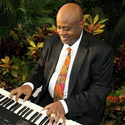 A man in a black suit plays a keyboard surrounded by lush greenery, appearing focused and content.