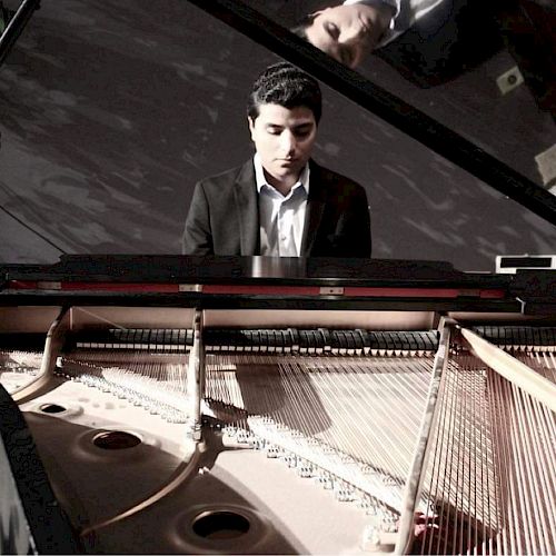 A person is playing a grand piano with the lid open, revealing the strings and hammers inside. The individual's reflection is visible in the lid.