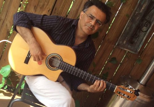 A person sitting on a chair playing an acoustic guitar, with a wooden fence and greenery in the background. The individual appears to be engaged in music.