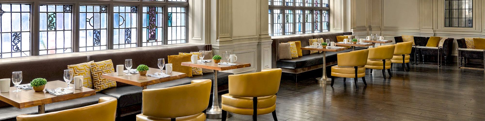 Elegant dining room with large arched windows, yellow chairs, long wooden tables set with plates, and crystal chandeliers overhead.